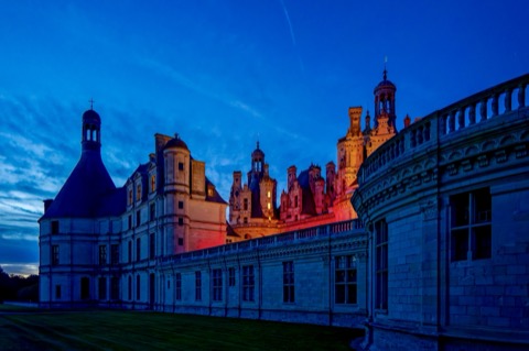 Les toits et tourelles Renaissance du château de Chambord se découpent sur un ciel bleu profond à la tombée de la nuit, certaines façades éclairées d'une lumière rouge-orangé, vues depuis la terrasse.