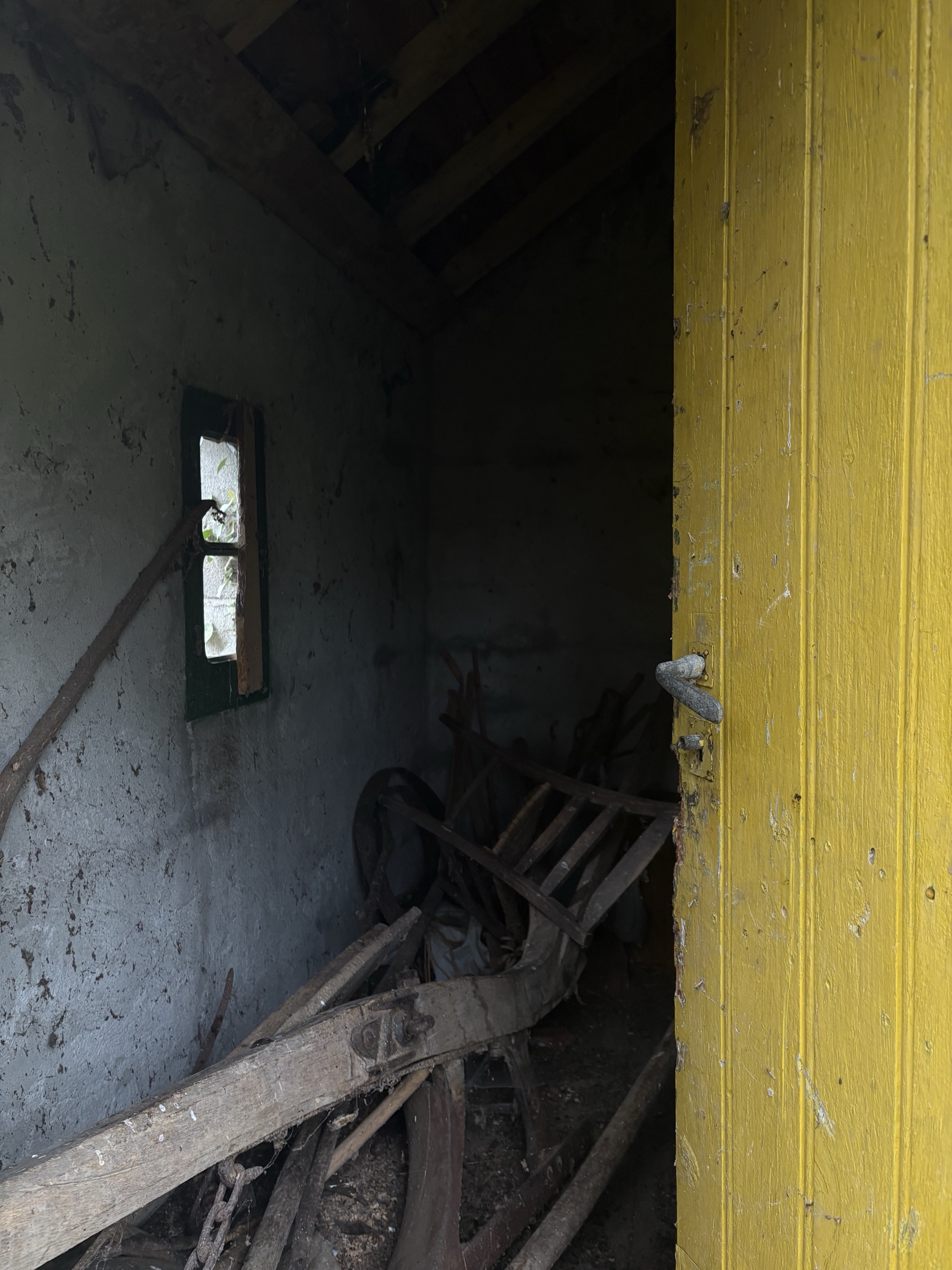 Une porte en bois peinte en jaune vif, entrouverte sur une remise sombre : sous une charpente apparente, du vieux matériel agricole en bois est entassé contre un mur gris-bleu, éclairé par une petite fenêtre étroite.