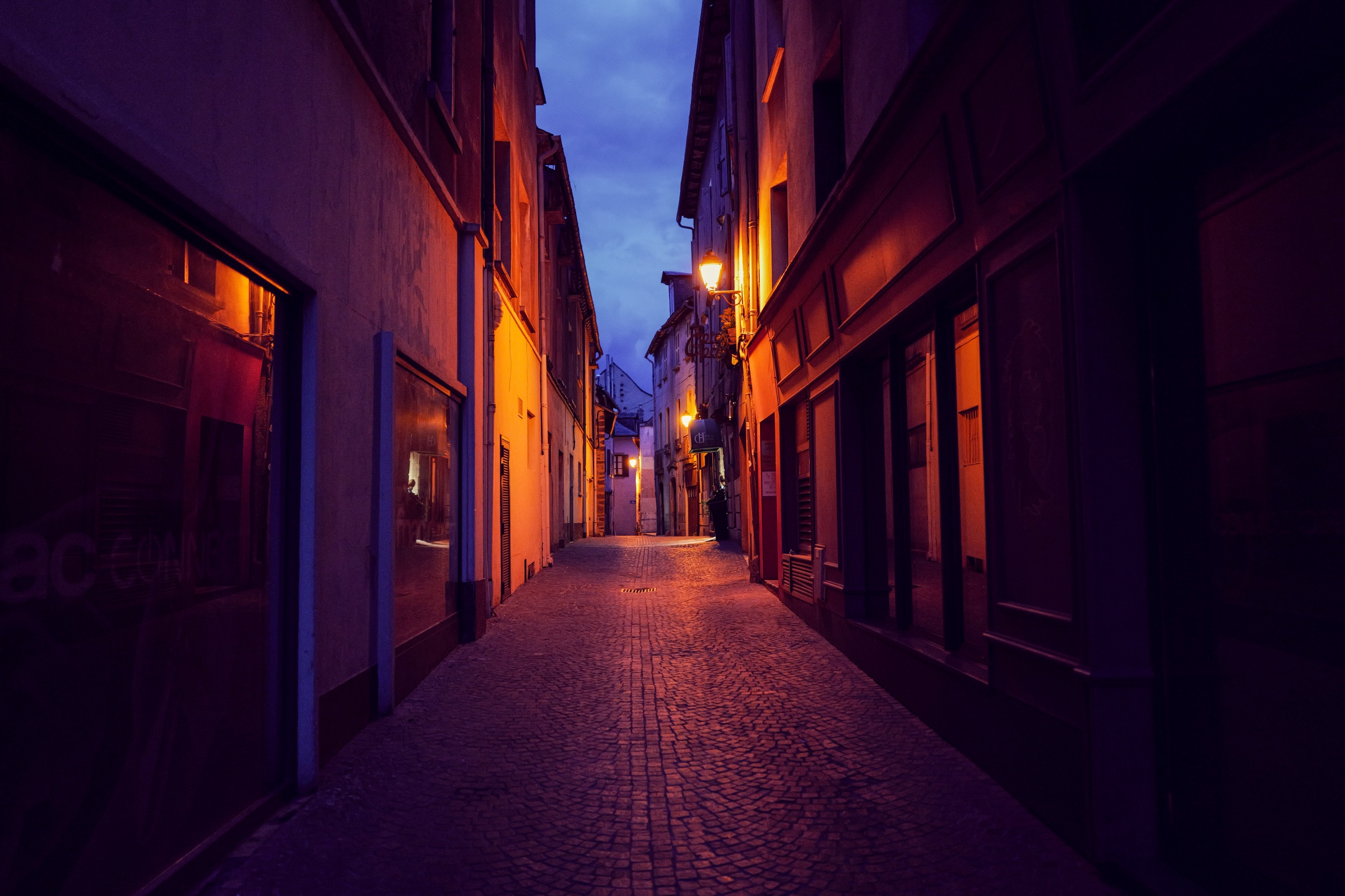 Une ruelle pavée et étroite de Rodez à la tombée de la nuit, éclairée par des réverbères à la lumière chaude, entre des façades sombres, sous un ciel bleu profond.