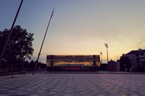 Façade rectangulaire et vitrée de la salle des fêtes de Rodez au crépuscule, semée de points lumineux en LED évoquant la voie lactée, sur une place pavée et déserte.