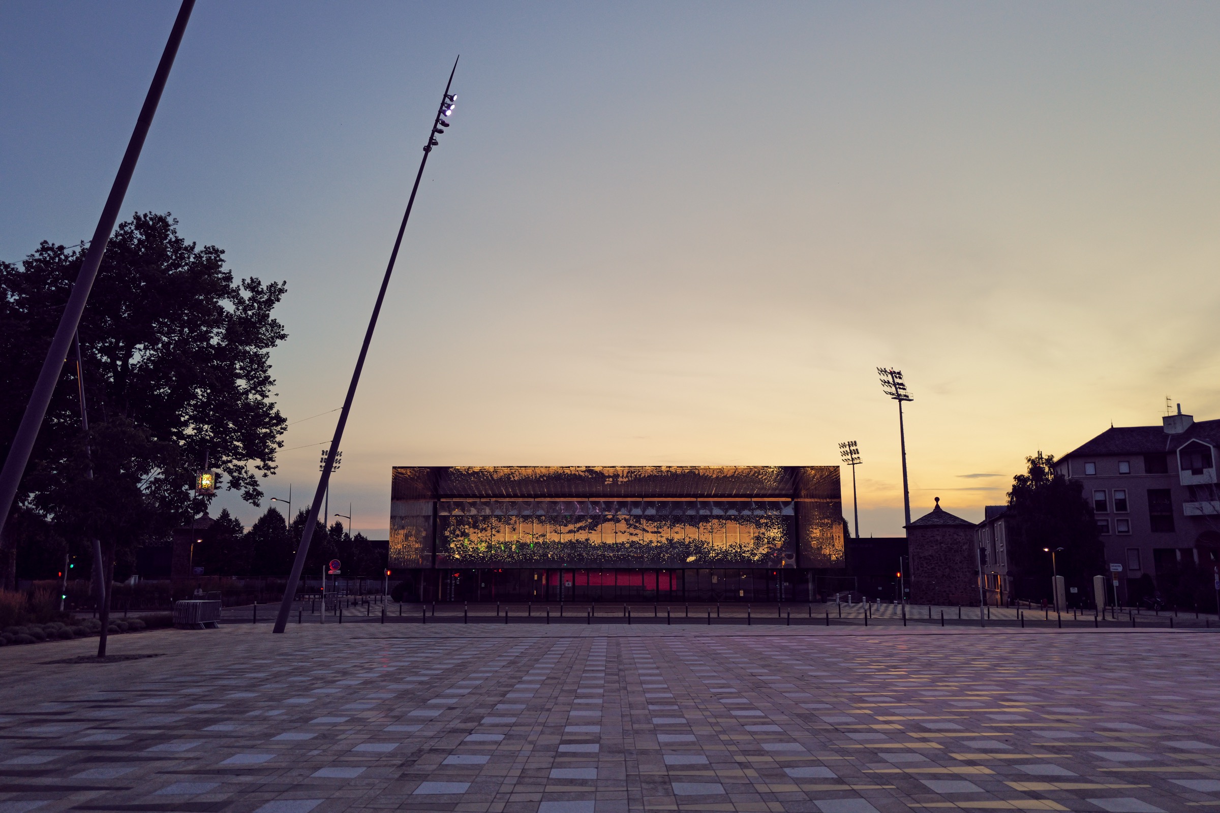 Façade rectangulaire et vitrée de la salle des fêtes de Rodez au crépuscule, semée de points lumineux en LED évoquant la voie lactée, sur une place pavée et déserte.